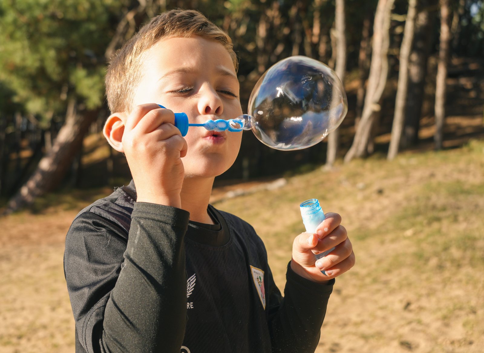 A boy blowing a big soap bubble among pine trees on a sunny day
